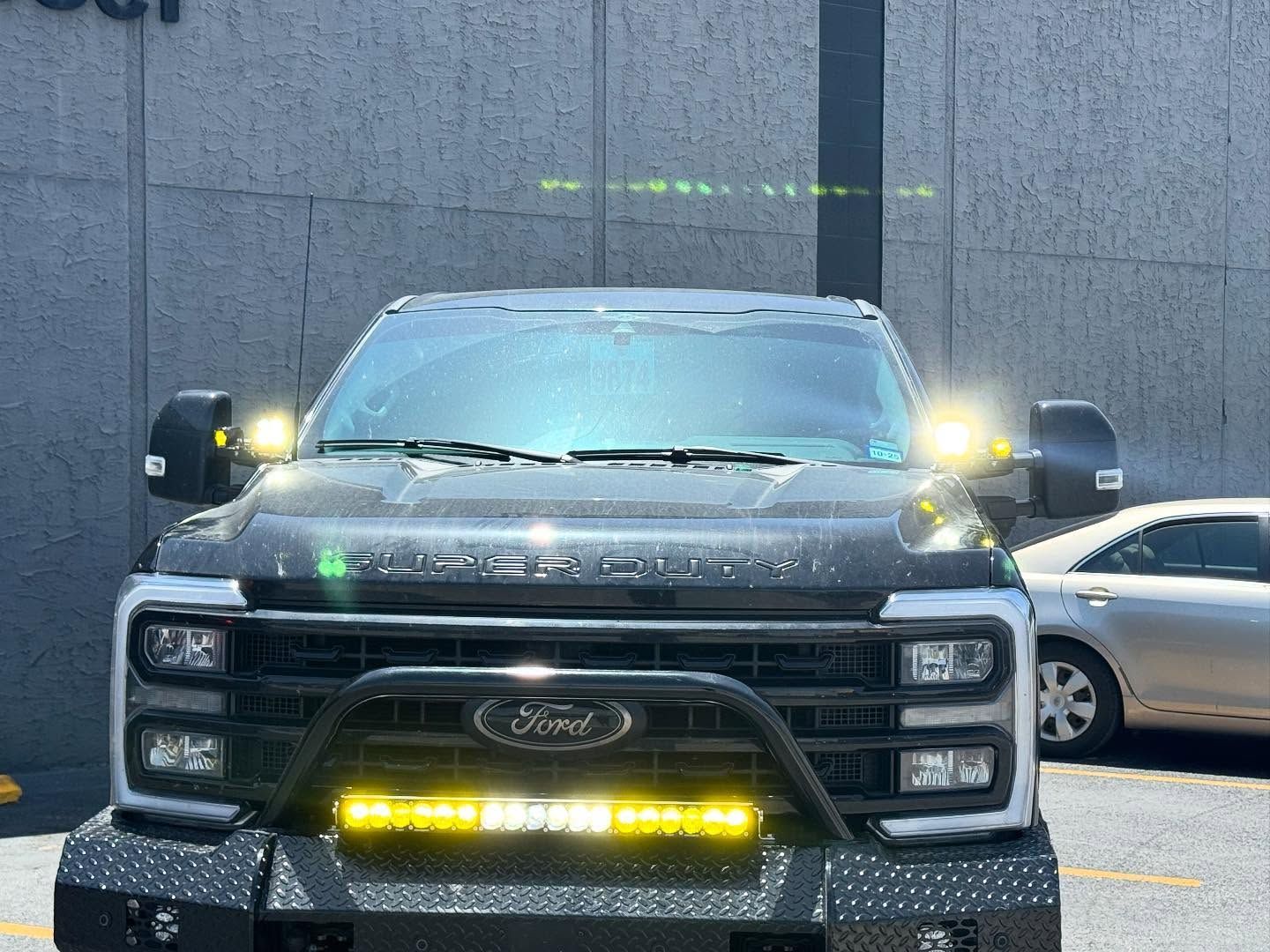 Black Ford pickup with bright lights parked in front of a gray wall and a white car behind it.