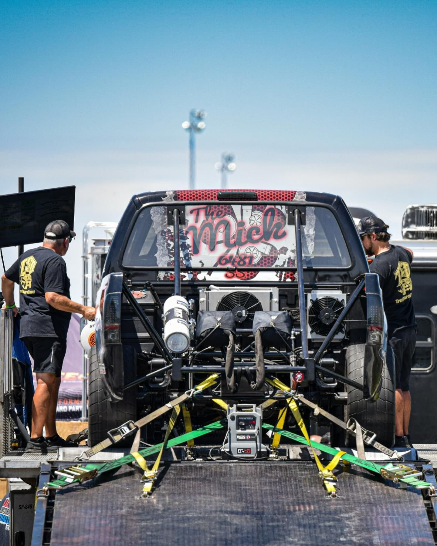 Rear view of a race truck with crew members attaching green and yellow tow straps in a pit area