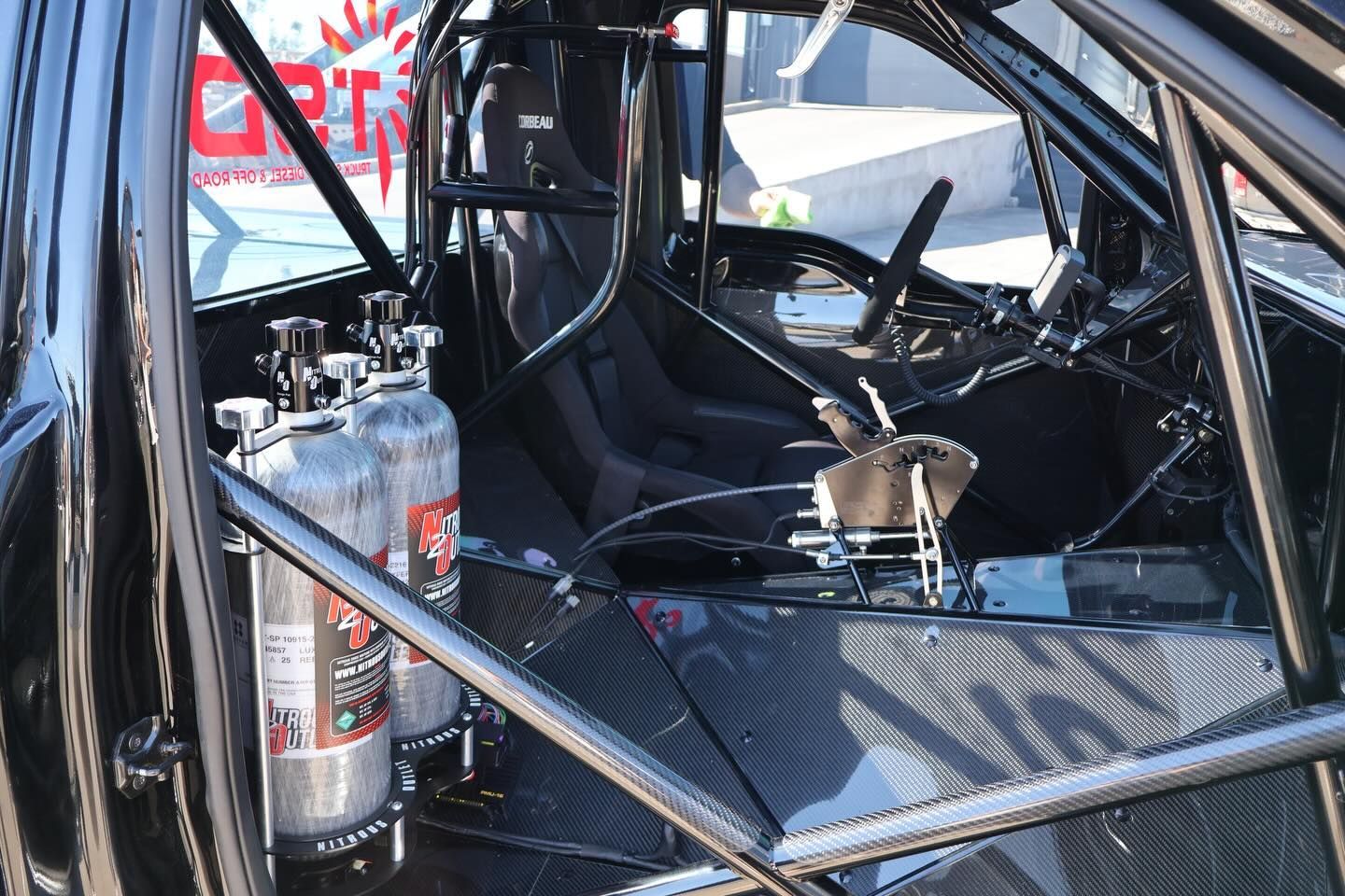 Interior of a race car with a roll cage, racing seat, fire extinguisher, and carbon-fiber floor panel.