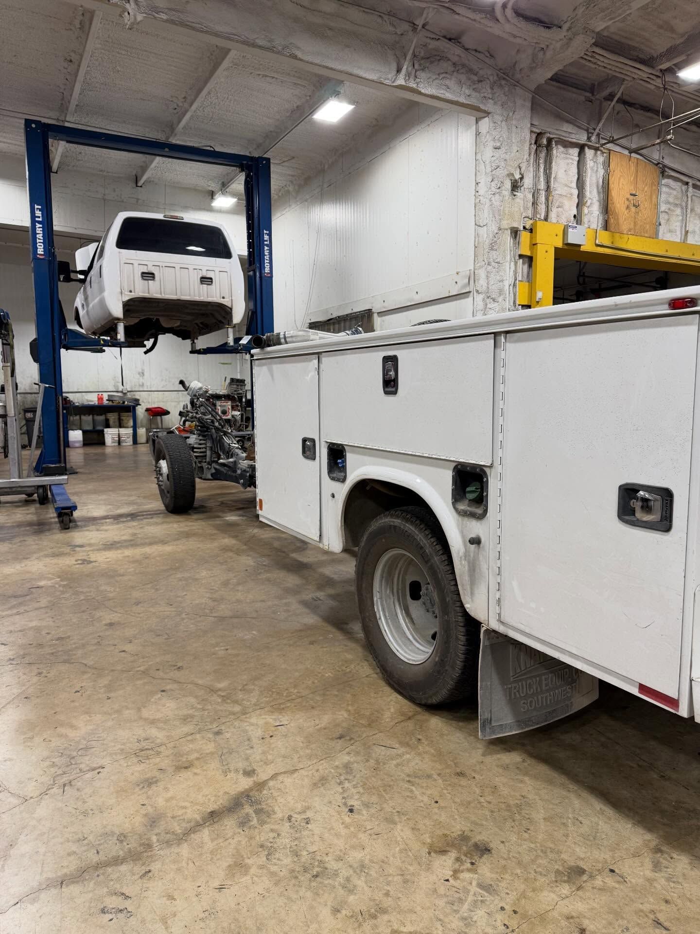 White vehicle lifted in a garage beside a white utility truck and blue repair lift.