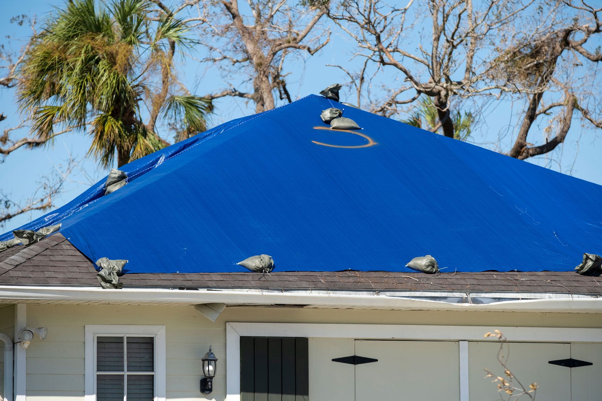 Blue tarp covering damaged roof of a light-colored house.