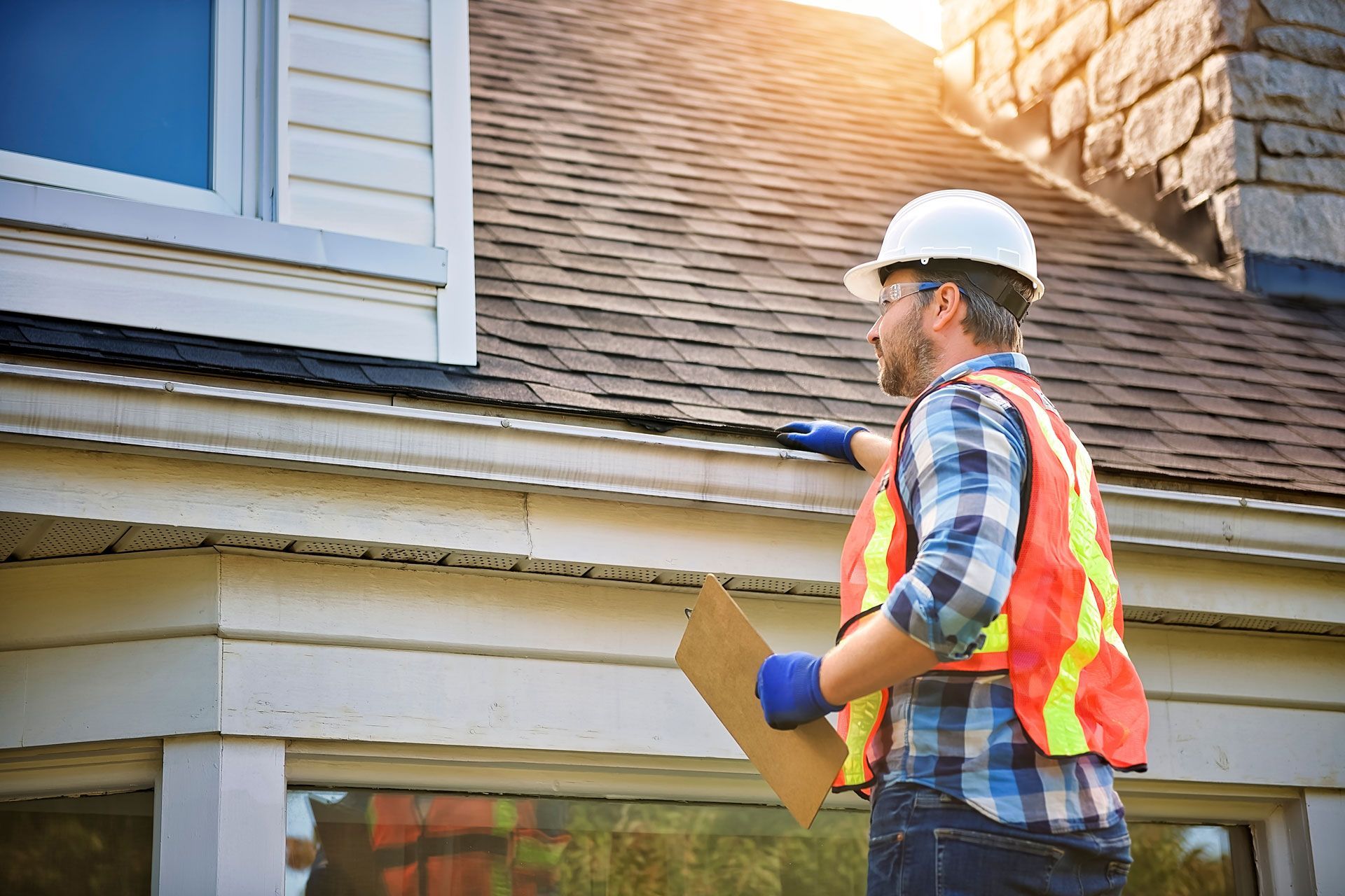Man in hard hat and safety vest inspecting a house's gutter.