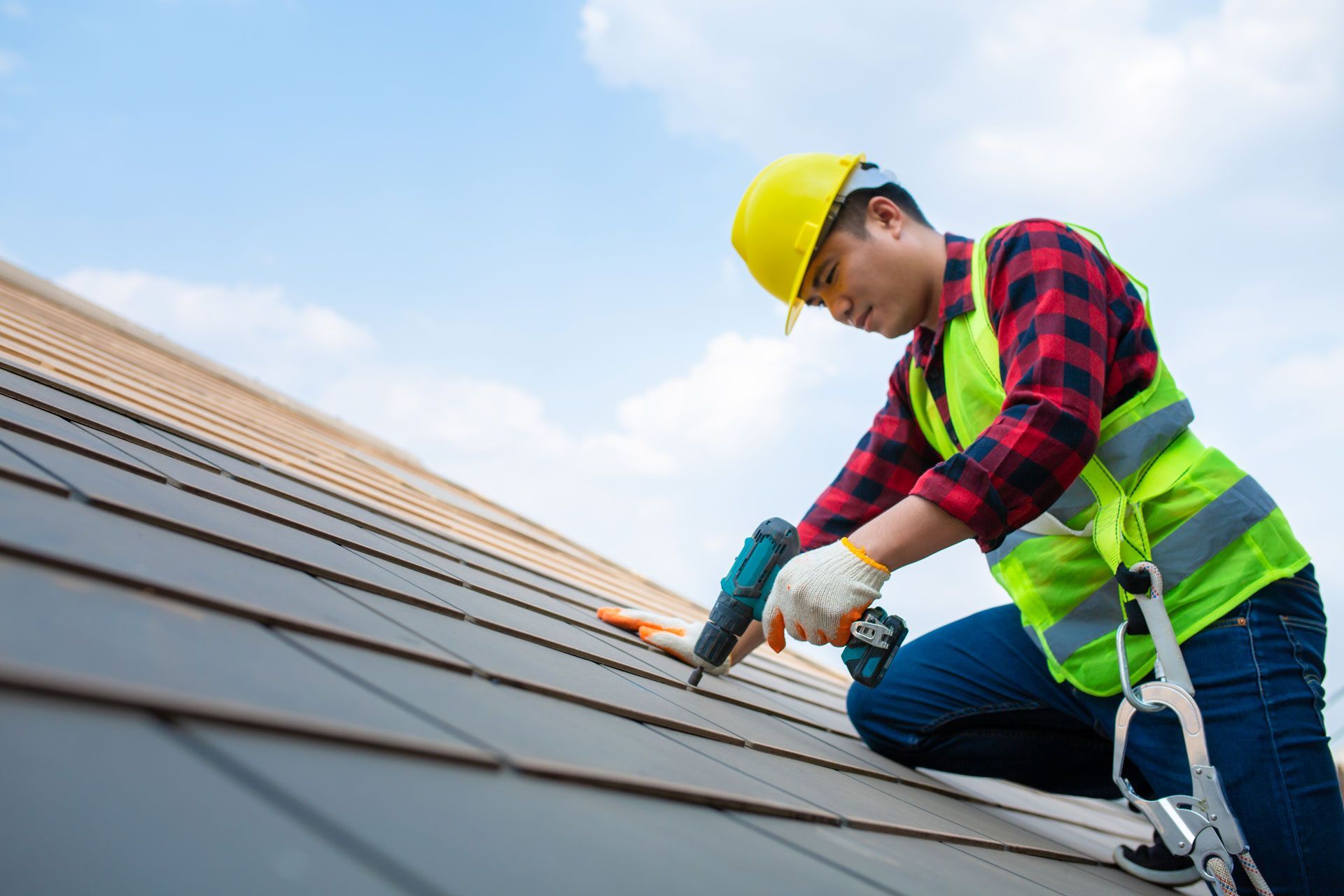 Roofer in hard hat and safety vest using a caulking gun on a tiled roof under a blue sky.