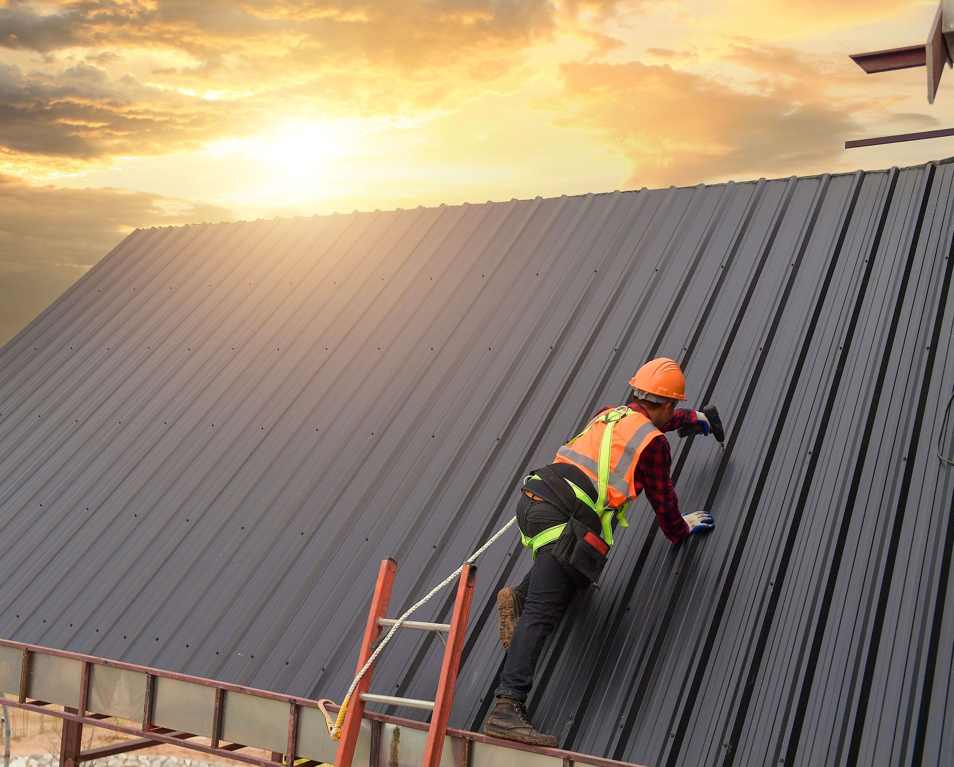Roofer in safety gear on a metal roof, securing material. Ladder present, sunset backdrop.