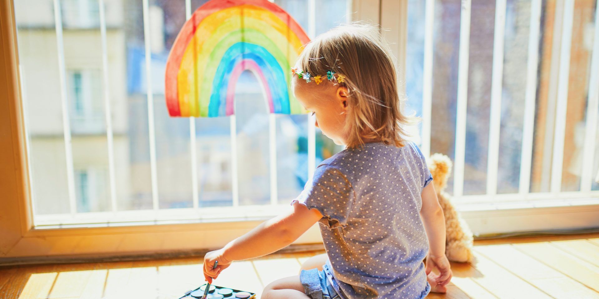 A little girl is sitting on the floor looking at a rainbow painted on a window.