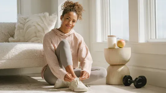 Woman sitting on the floor, tying her shoelace. She wears a pink sweater, gray leggings.