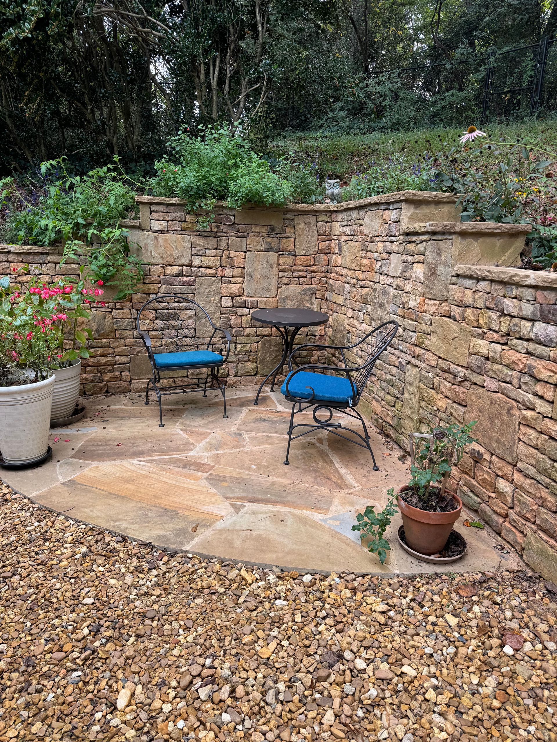 Small stone patio with two blue chairs, round table, potted plants, and a brick wall in a garden.