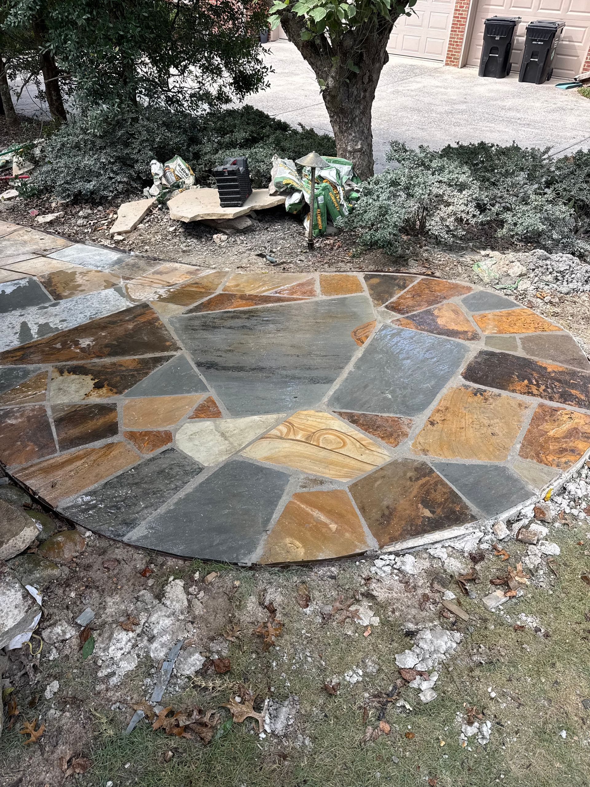 Stone patio with a circular mosaic pattern beneath a tree, surrounded by mulch and grass.