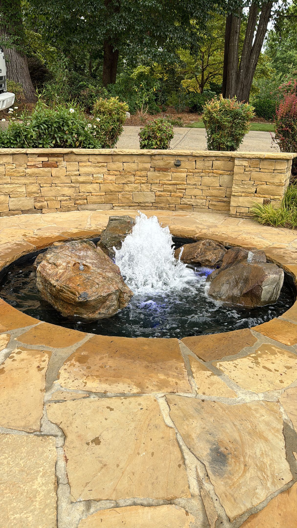 Small stone garden fountain with water bubbling between two rocks, set against a low wall and greenery.