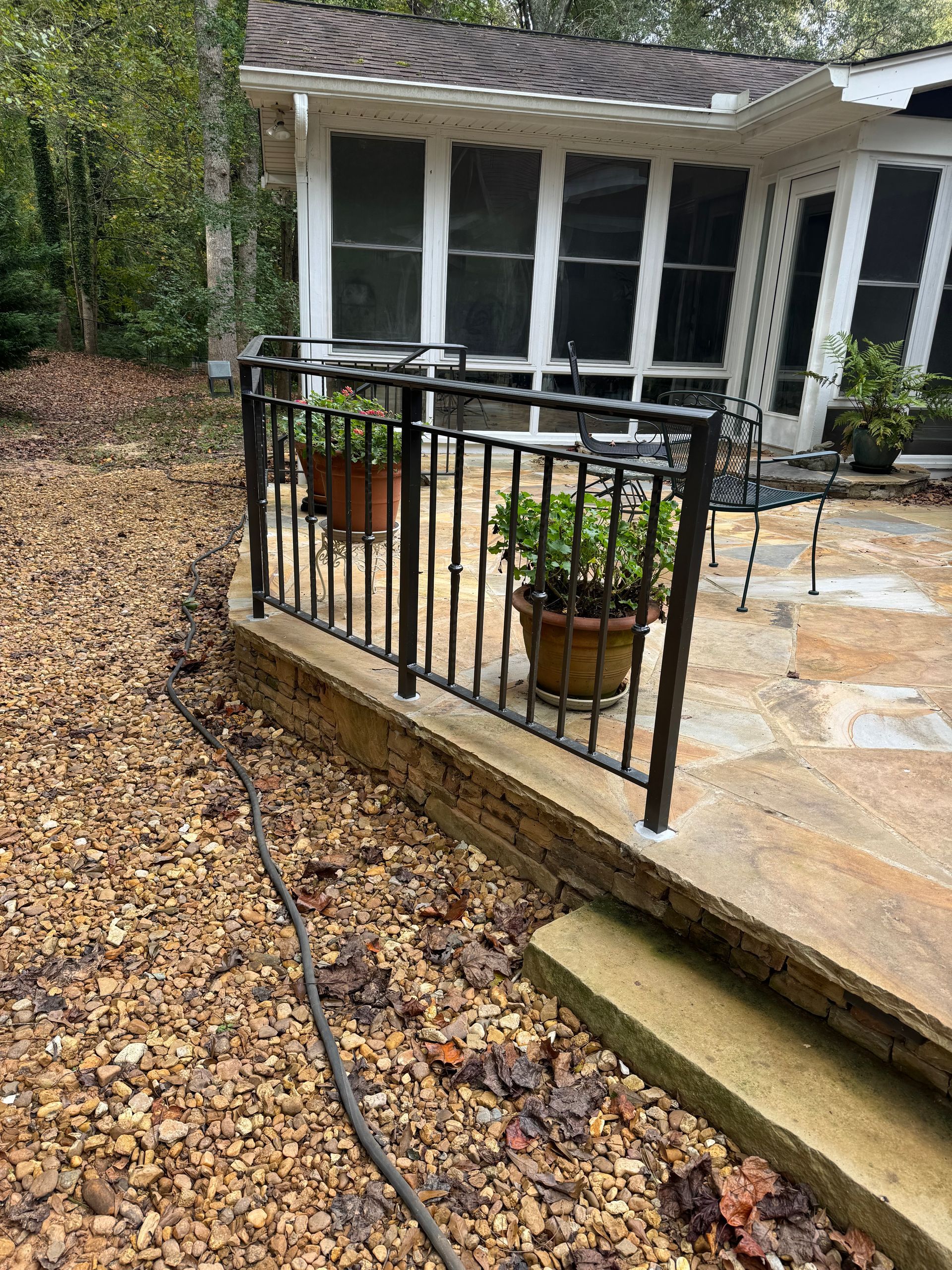 Back patio with black metal railing, potted plants, and a hose on a leaf-covered gravel path.