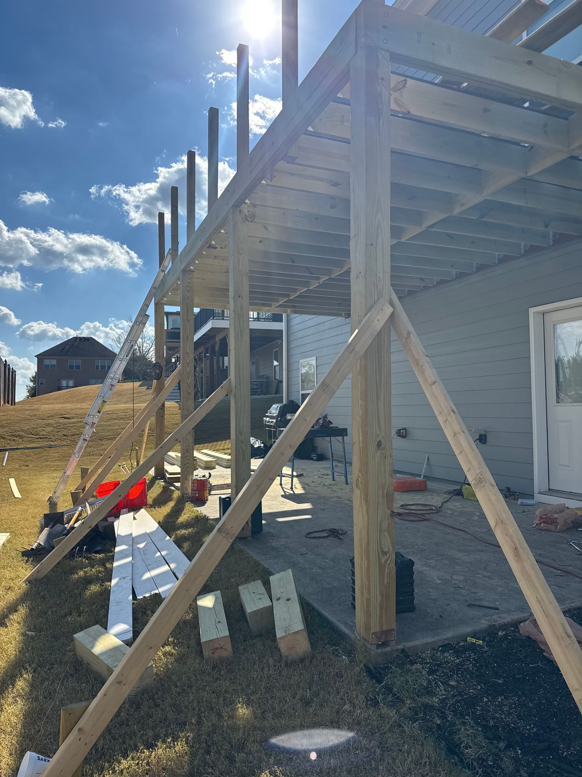 Wooden deck framing under construction beside a gray house, with stairs and supports in bright sunlight.