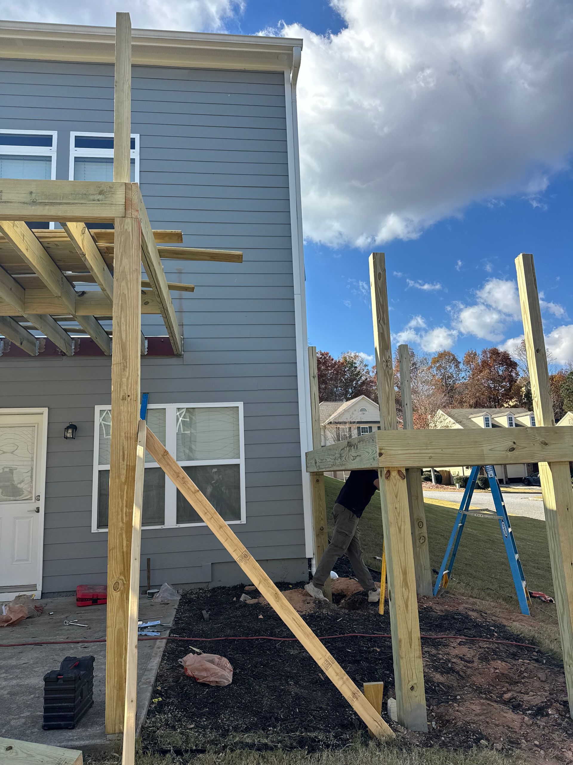 Partially built wooden deck beside a gray house, with support posts and beams under a cloudy sky.