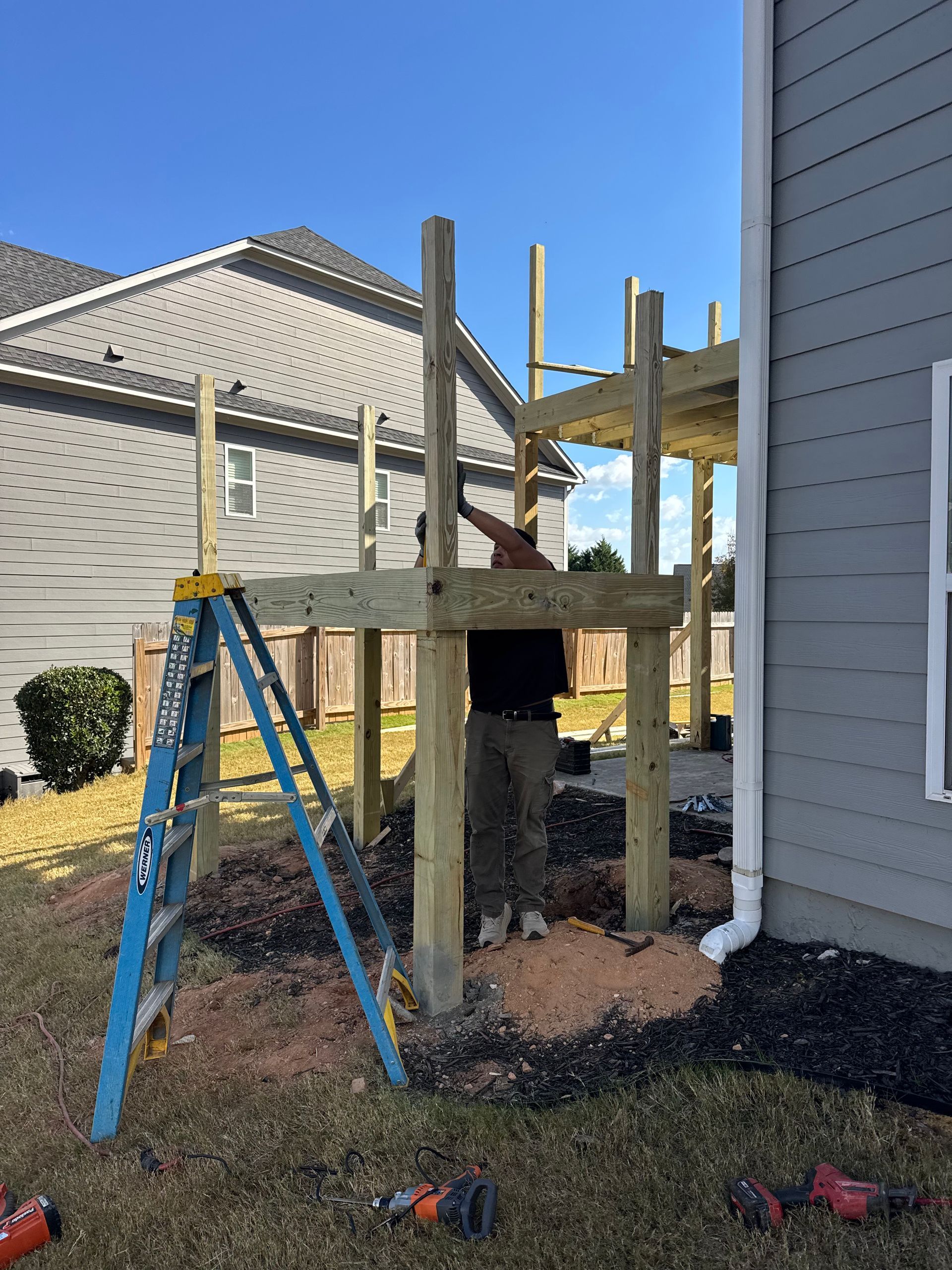 Wooden deck under construction beside a house, with ladder, posts, and scattered tools in a yard.