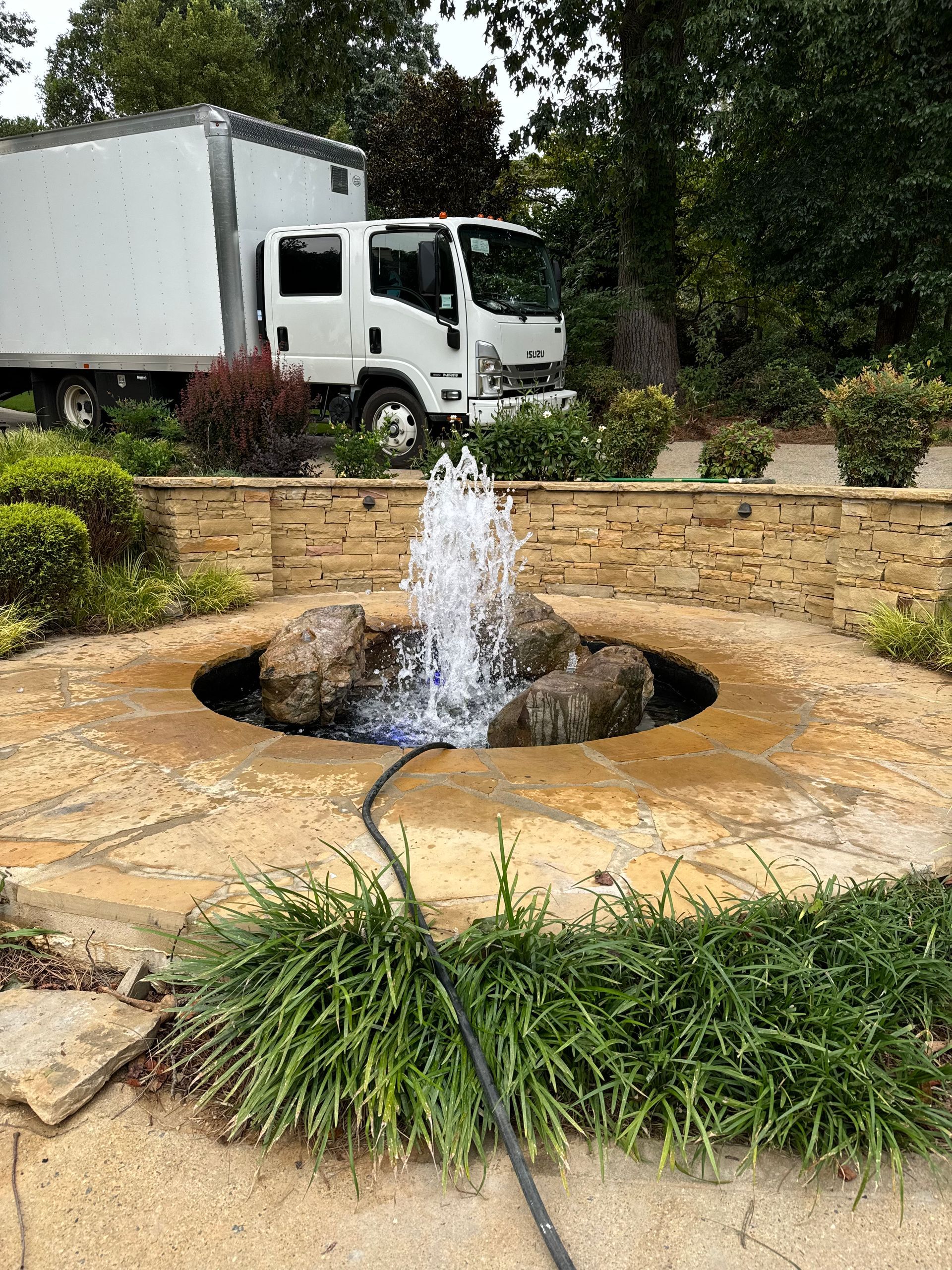 Stone fountain spraying water in a landscaped courtyard, with a white truck and trees in the background