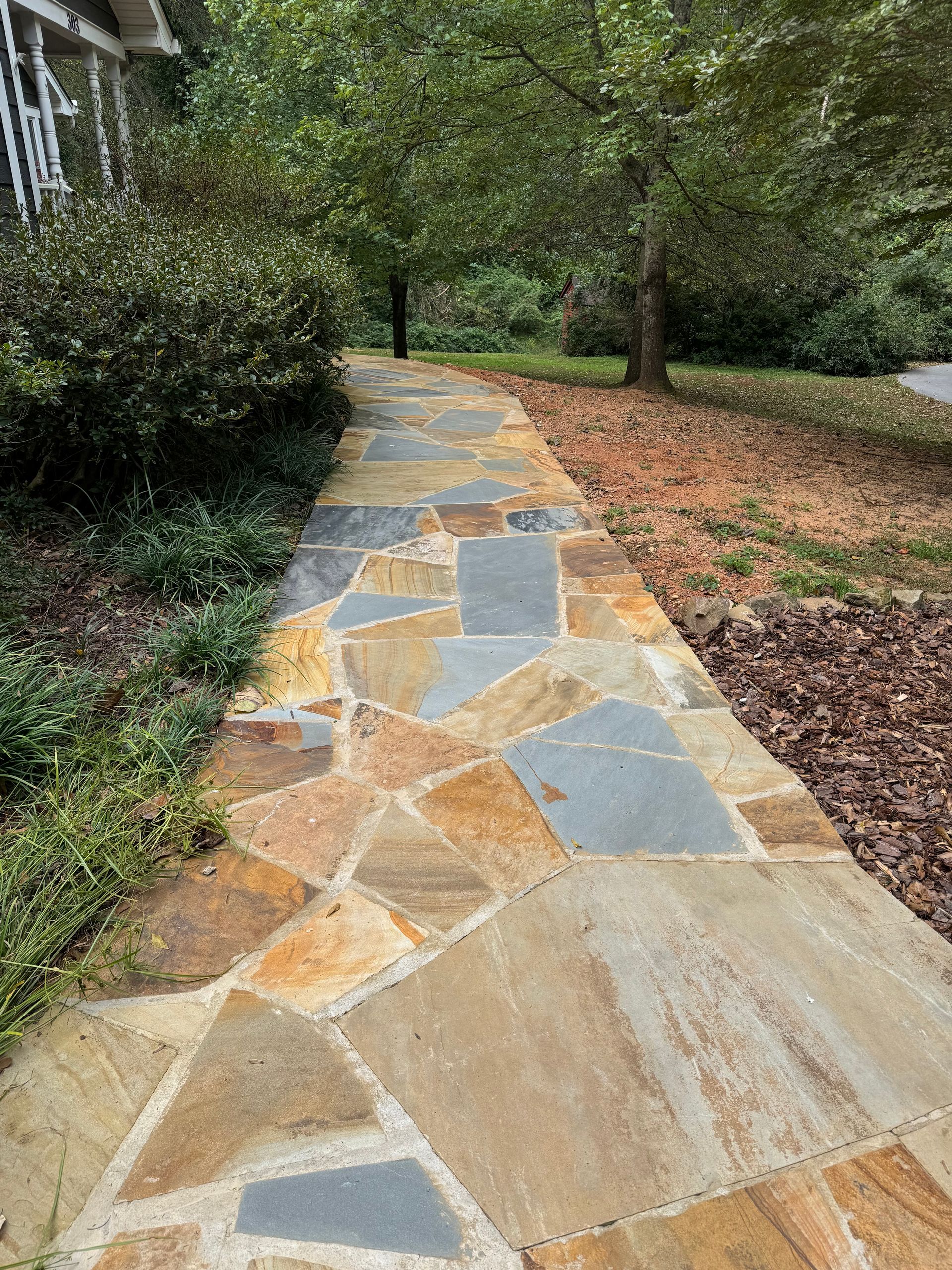 Stone walkway through a green garden with trees and shrubs, bordered by mulch and grass.