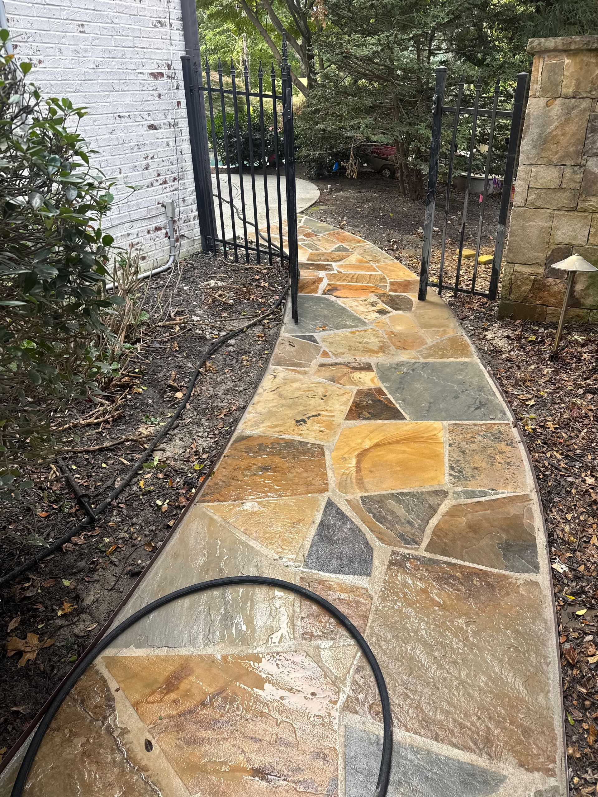 Curved stone garden walkway beside a white wall and iron gate, with mulch and greenery along the path