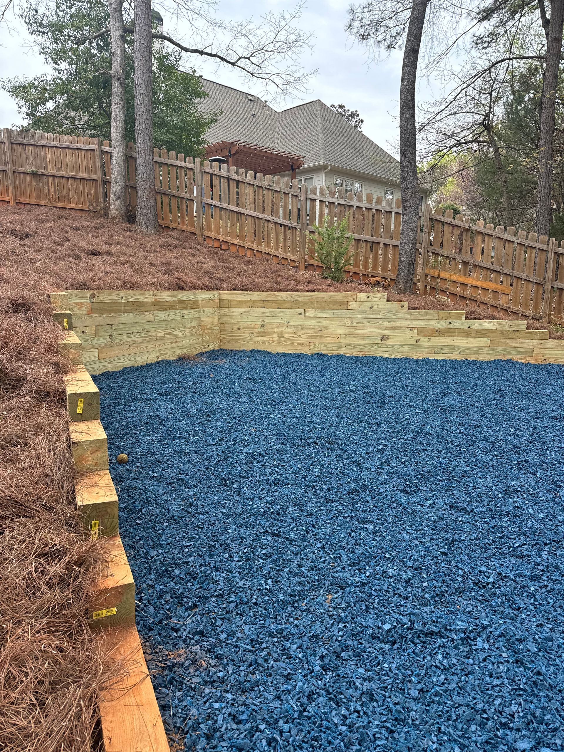 Backyard with a blue gravel area, dirt slope, retaining blocks, and a wooden fence under trees