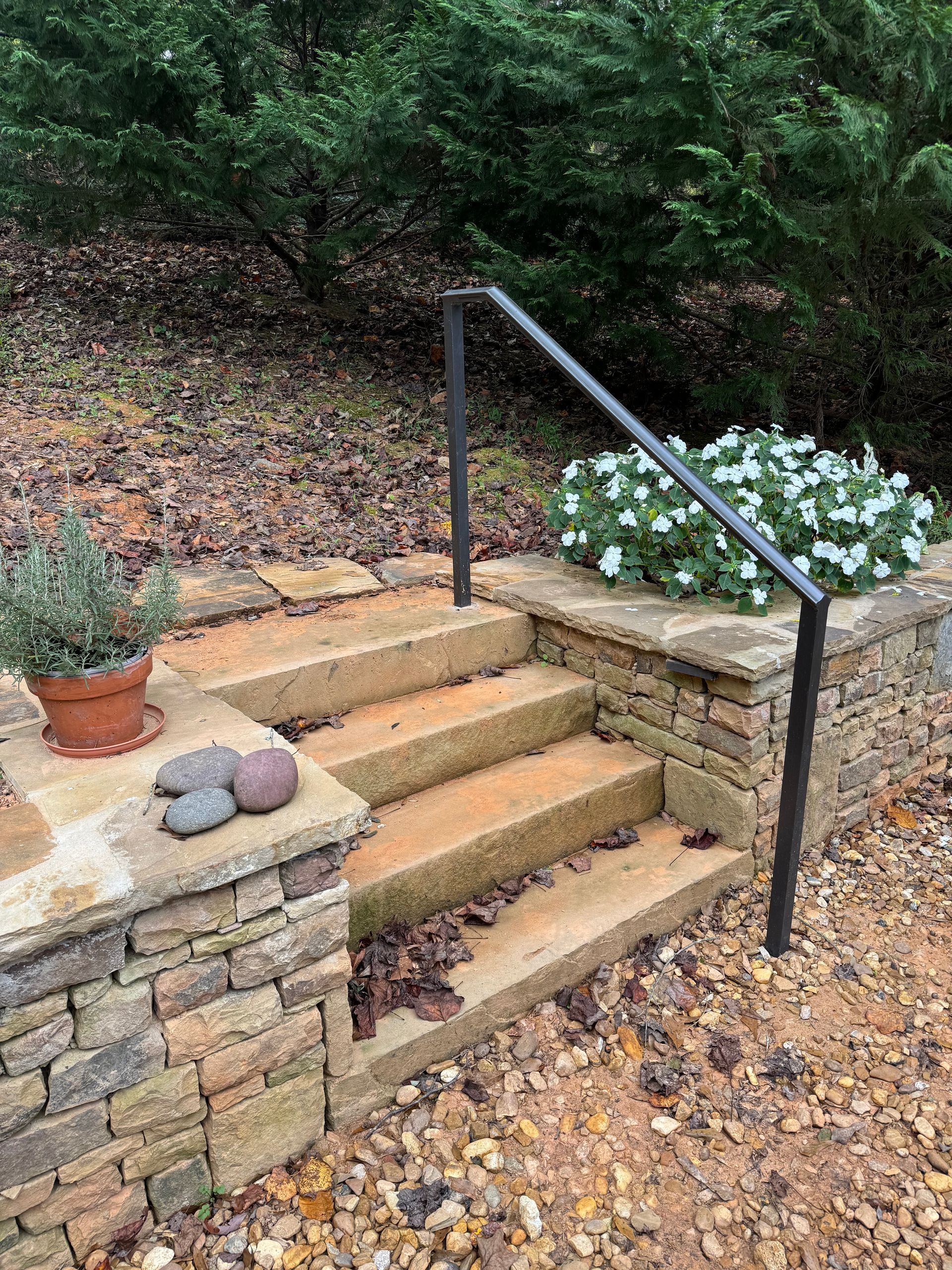 Stone steps with black handrail leading to a garden path, bordered by stone walls and white flowers