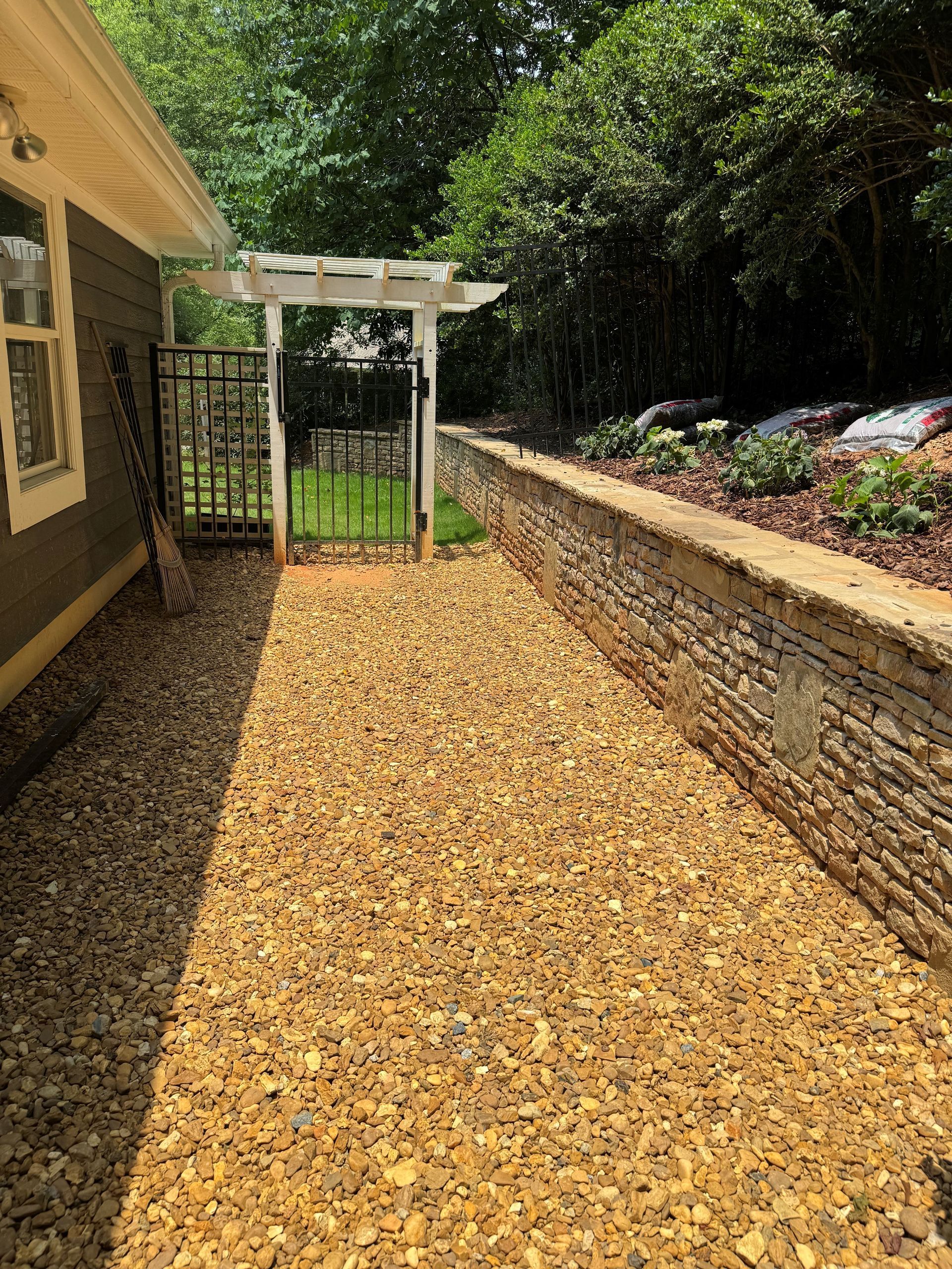 Gravel walkway beside a house, leading to a white pergola and garden bed with stone retaining wall.