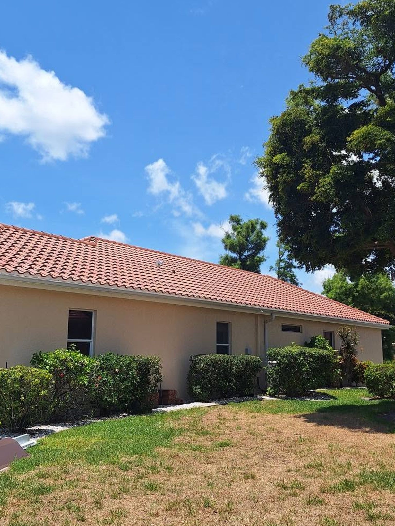 Single-story beige house with a red tile roof, shrubs, and a sunny green yard under a blue sky