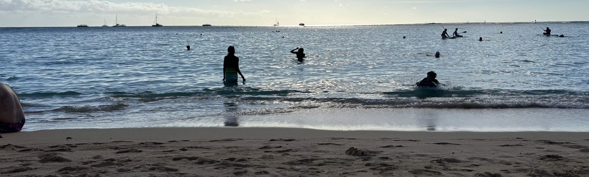 People wading and swimming at a calm beach under a cloudy sky, with waves and sand in the foreground