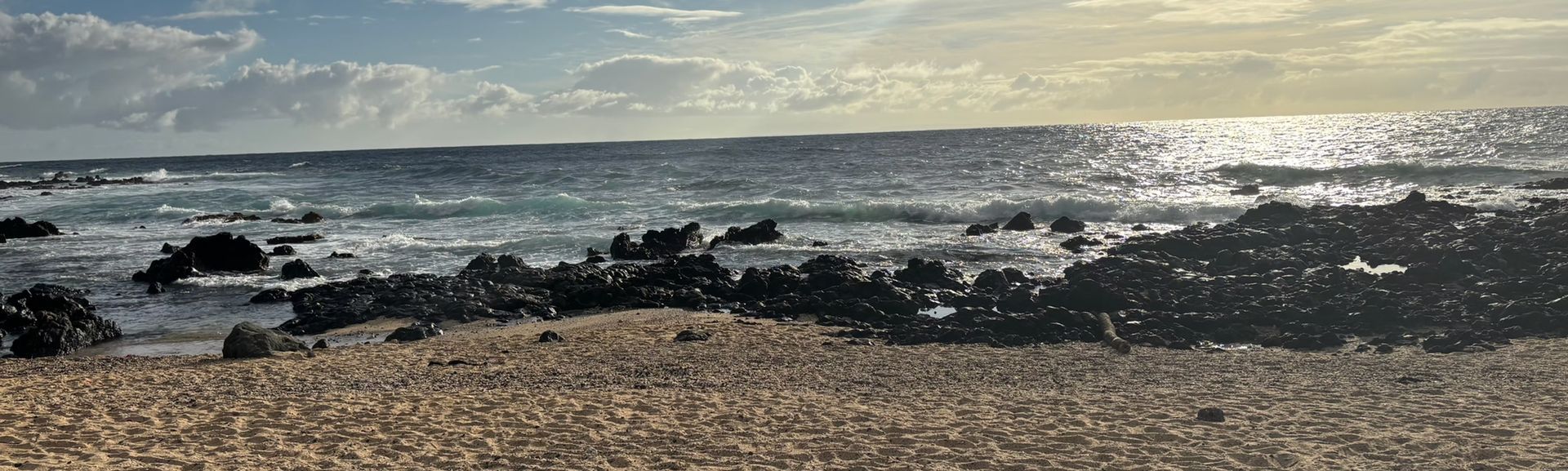 Rocky beach with waves, sunlit sea, and cloudy sky at sunset