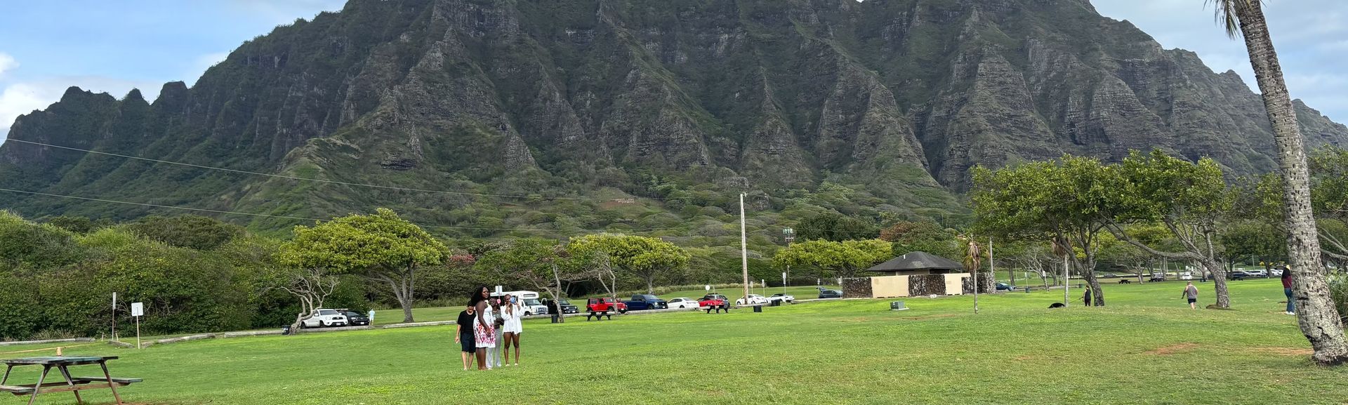 Wide grassy field with a few people and tents beneath a large rocky mountain under a cloudy sky