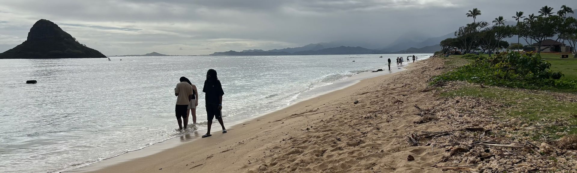 Two people walking along a sandy beach beside calm water under a cloudy sky