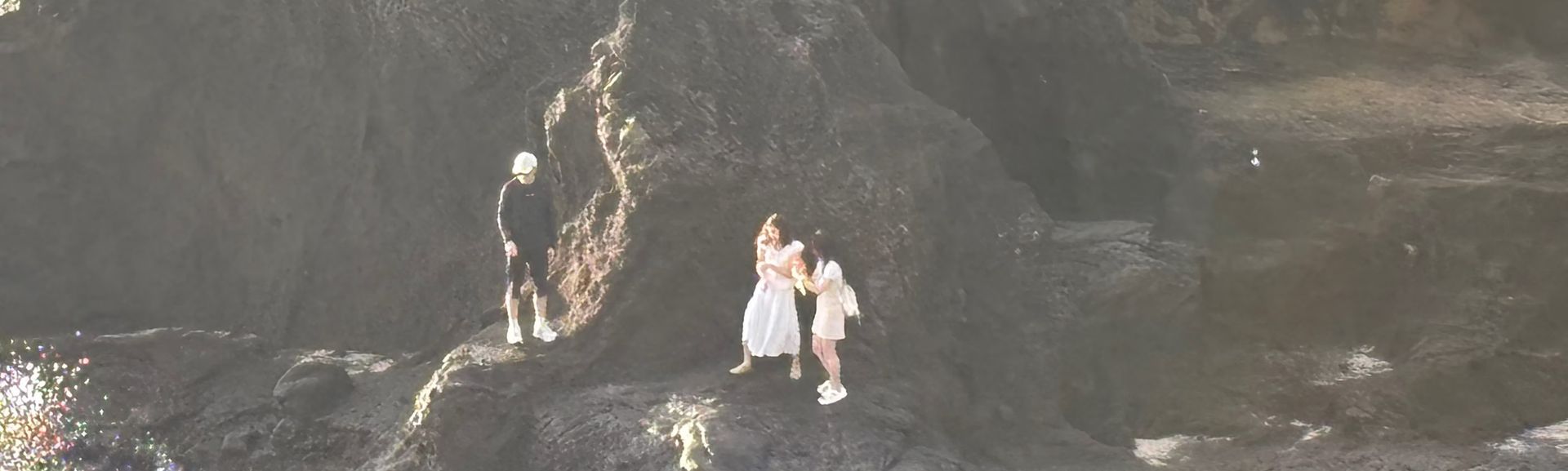Two people standing on dark rocky beach by a cliff and cave, with a small waterfall nearby