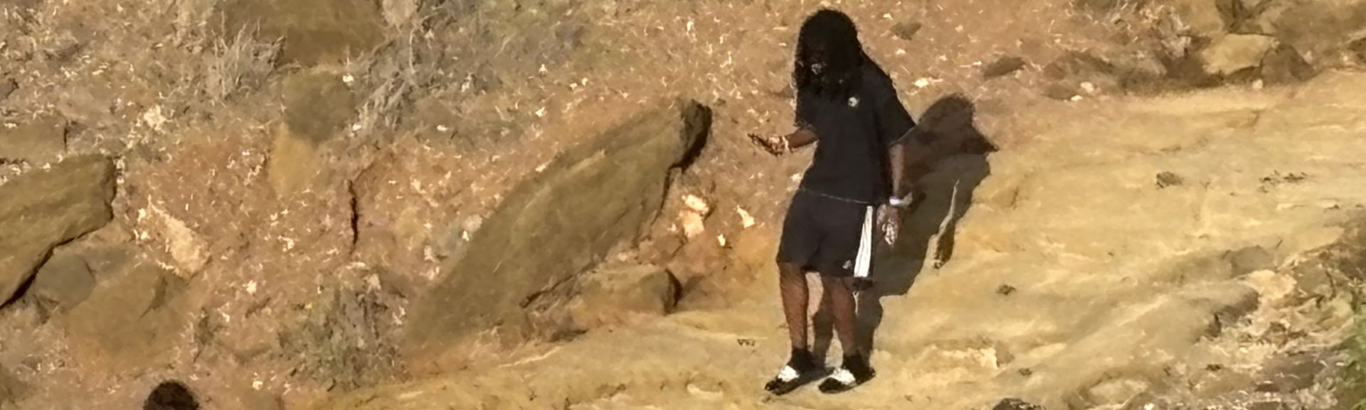 Person in black clothing standing on a rocky slope beside a large boulder.