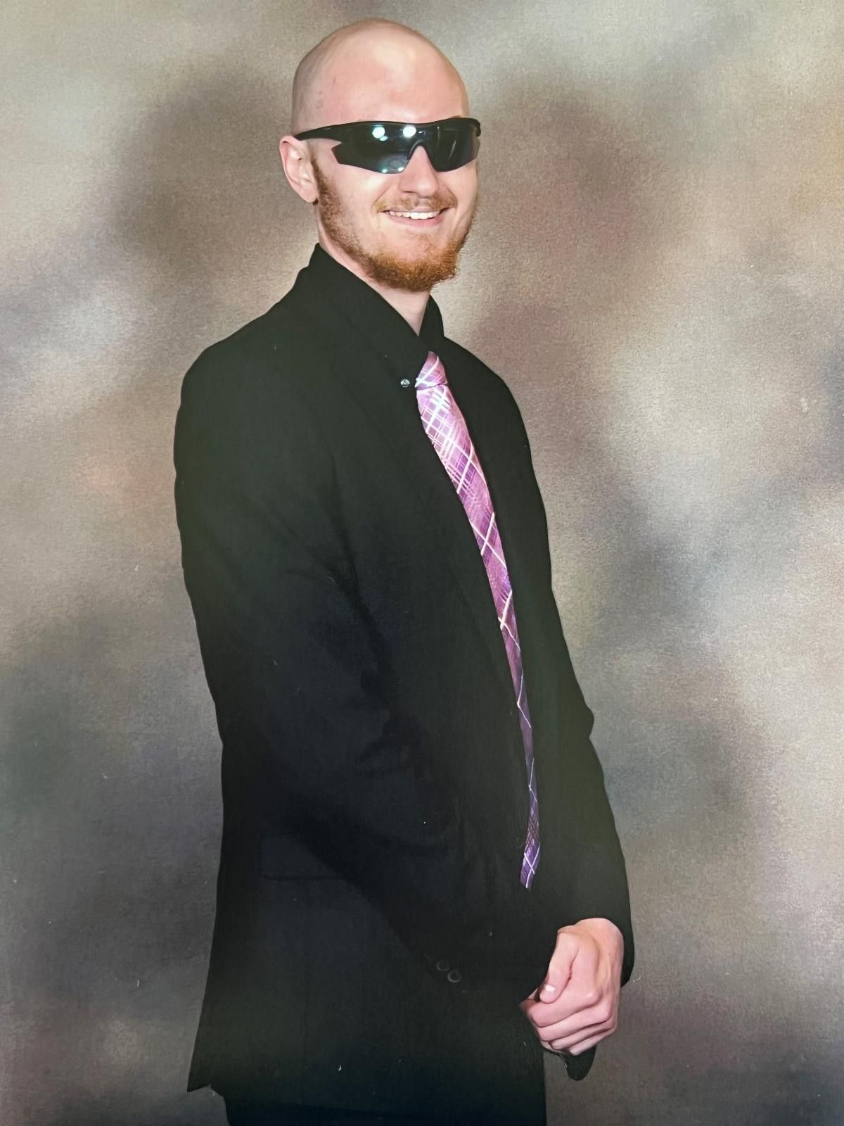 Bald man wearing sunglasses, black suit, and pink tie smiles, against a mottled backdrop.