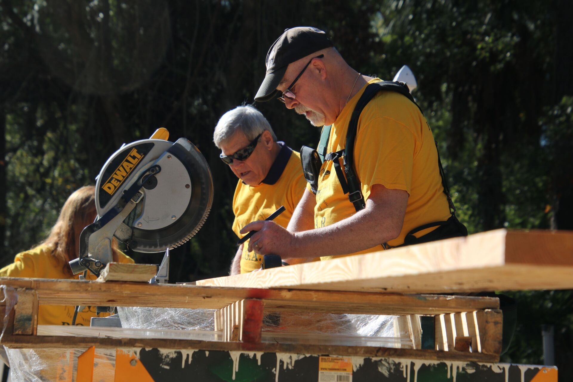 Two men in yellow shirts work with a saw and wood outdoors; a woman is partly visible.