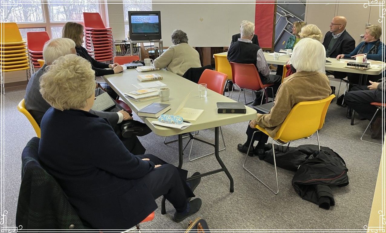 Group of older adults at a meeting around tables, watching a screen, some seated in yellow chairs.