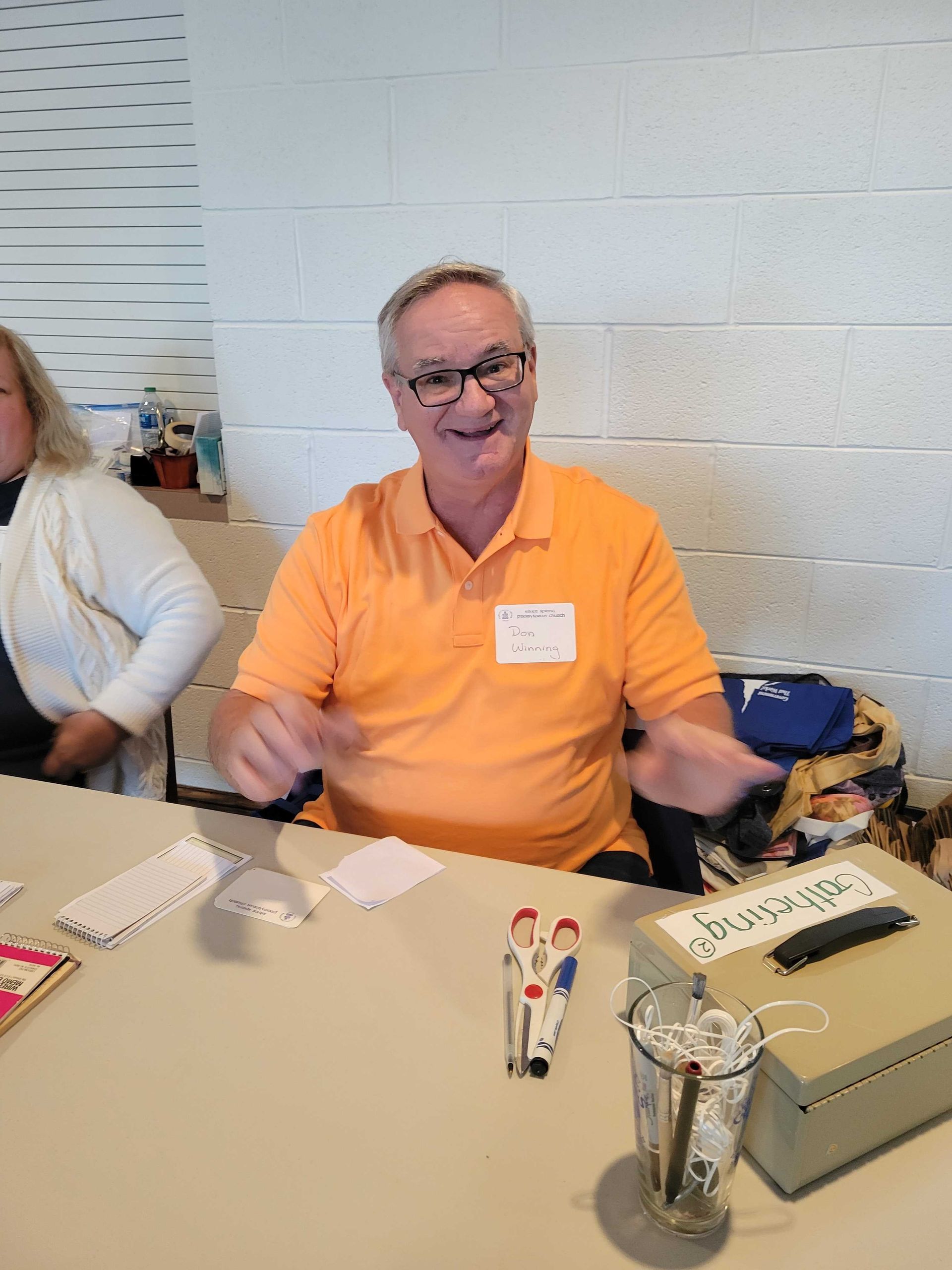 Man in orange shirt gestures, seated at table. Woman to left. Dominoes, cards, glass.