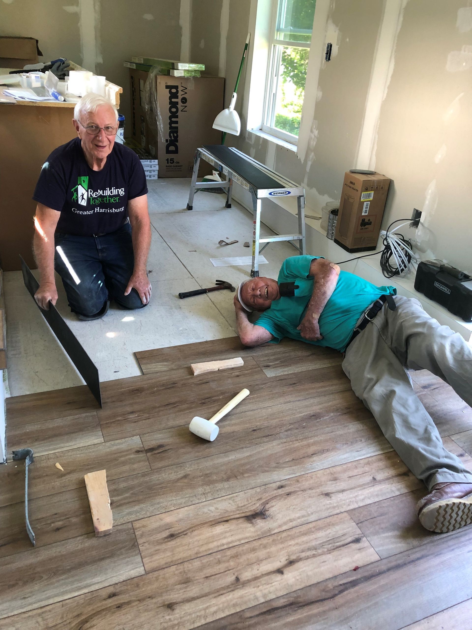 Two men installing wood-look flooring: One kneels, the other lies down. Hammer and tools are scattered on the floor.