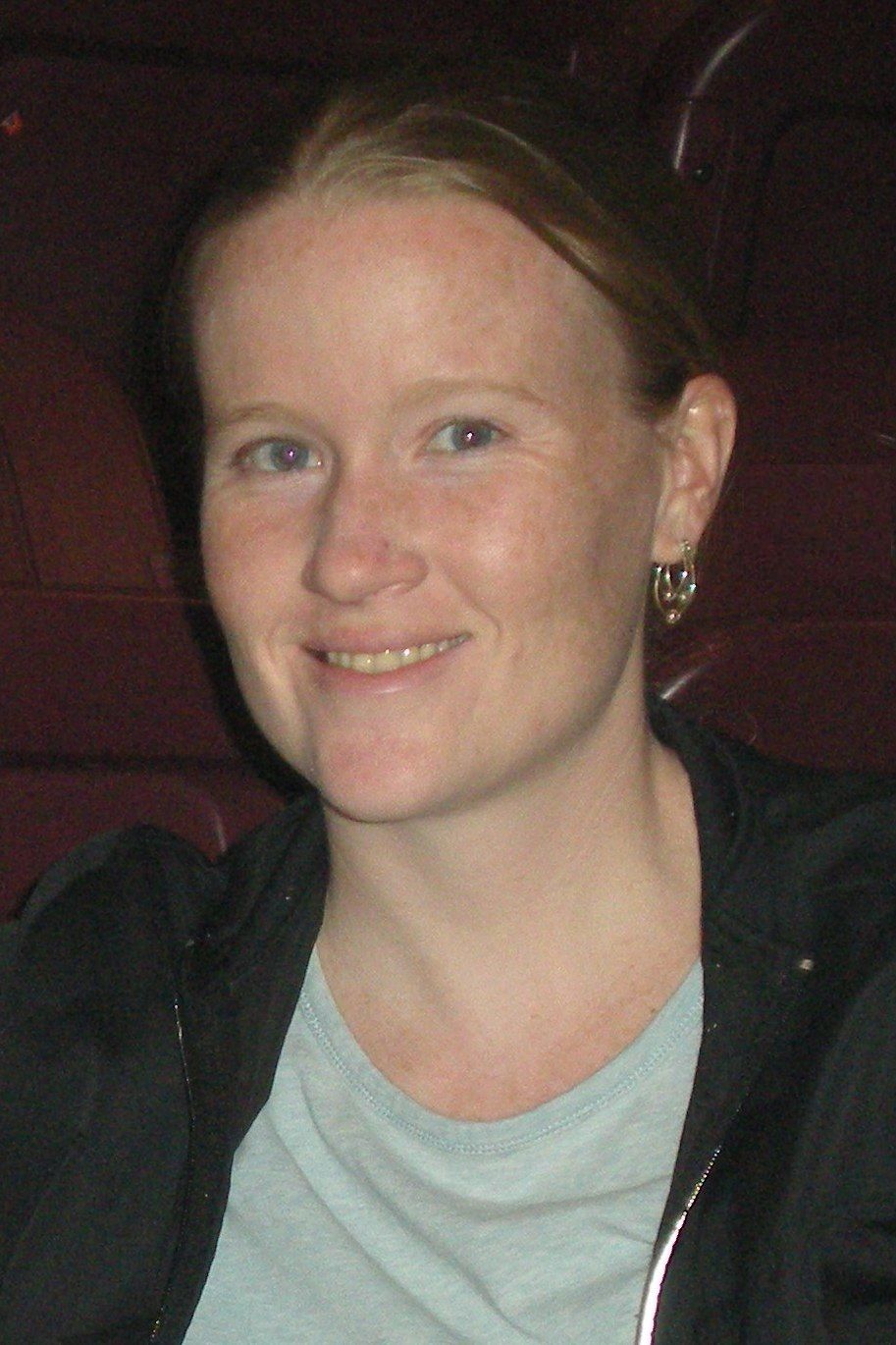 Woman with fair skin and red hair smiles, wearing a black jacket and a light blue shirt.