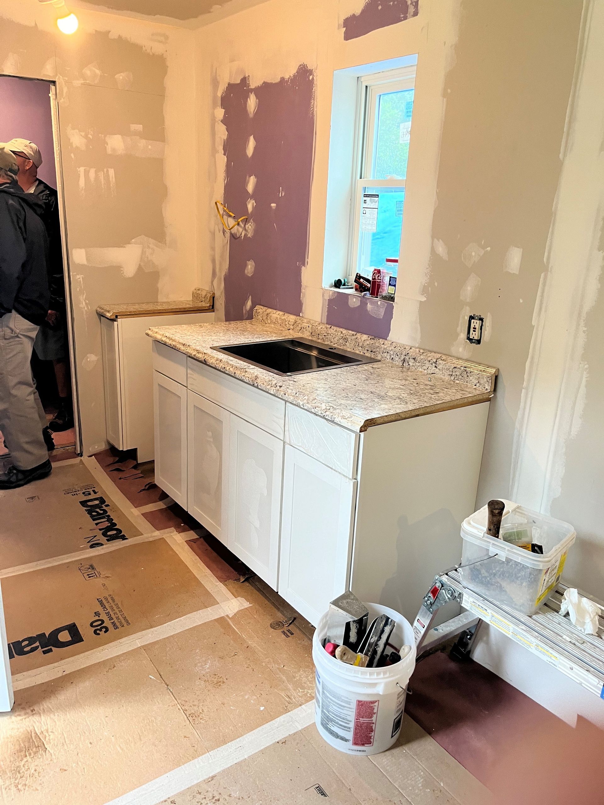 Kitchen renovation in progress with white cabinets, countertop, drywall, and a window. A person is visible on the left.