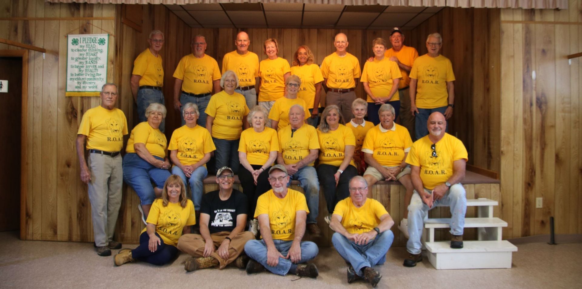 Group of people in yellow shirts posing indoors; wood paneling, some seated on steps.