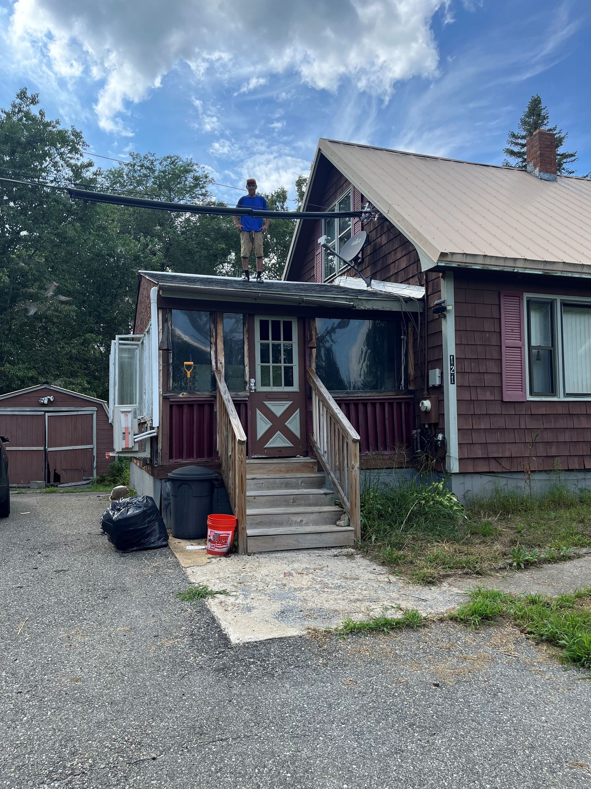 Person on a house roof with dark trim. Front porch with steps, gravel yard, and cloudy sky.