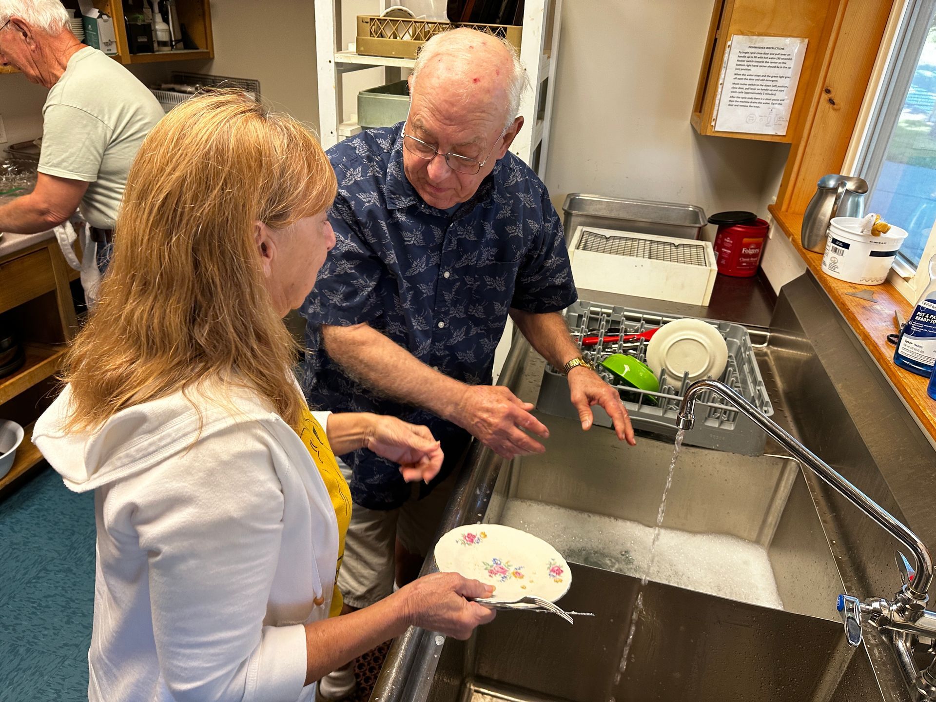 Woman and man wash dishes in a kitchen, talking. Another person is washing dishes at a separate sink.