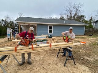 Two men cutting wood planks with saws, constructing a blue building outdoors.