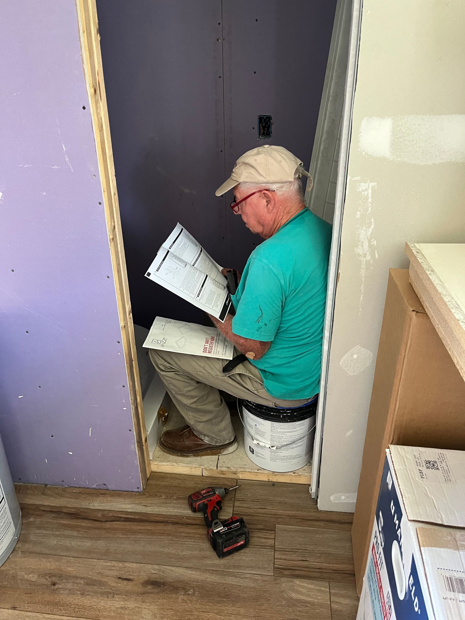 Man in a teal shirt and hat reads papers while sitting on a bucket in a small unfinished room.