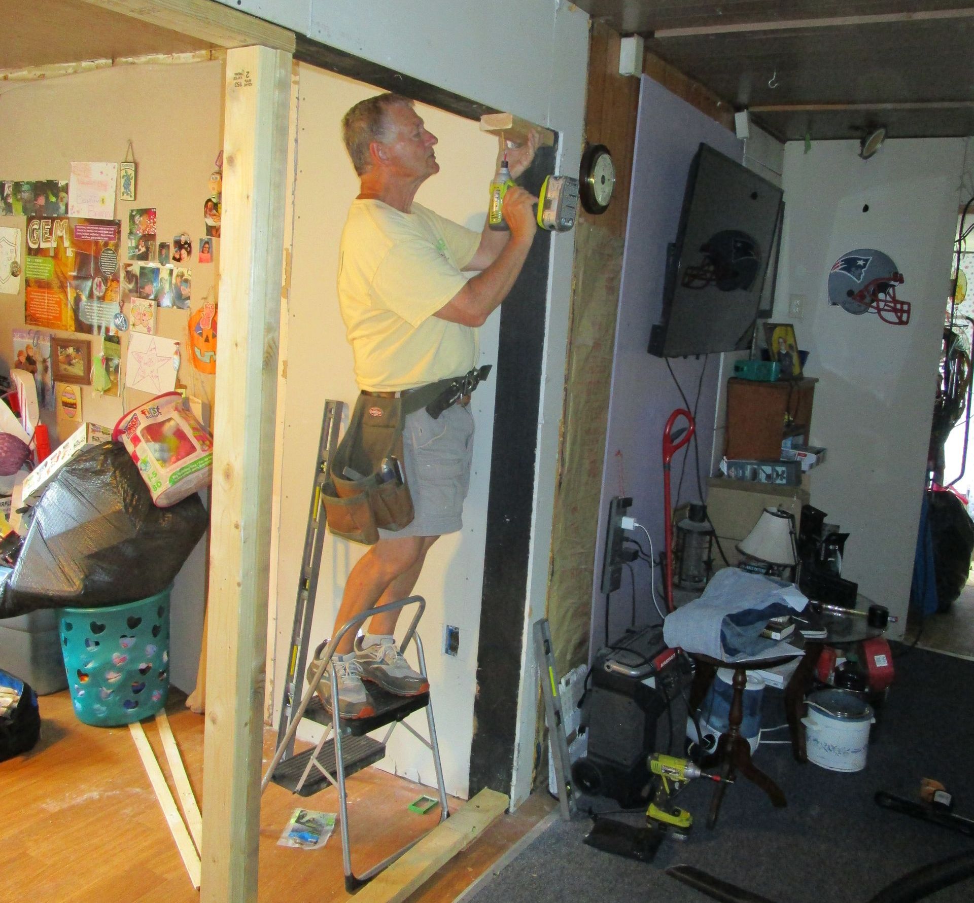 Man on stepladder installing a door frame; cluttered room with tools and decorations.