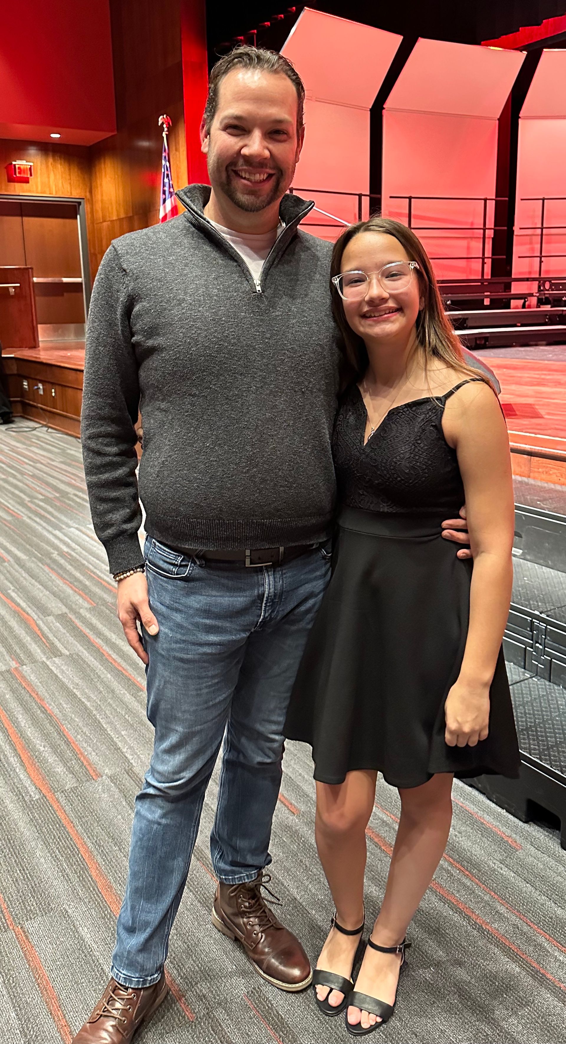 Man and girl pose together; man wears gray sweater and jeans, girl wears a black dress, both smile. Red-lit theater backdrop.