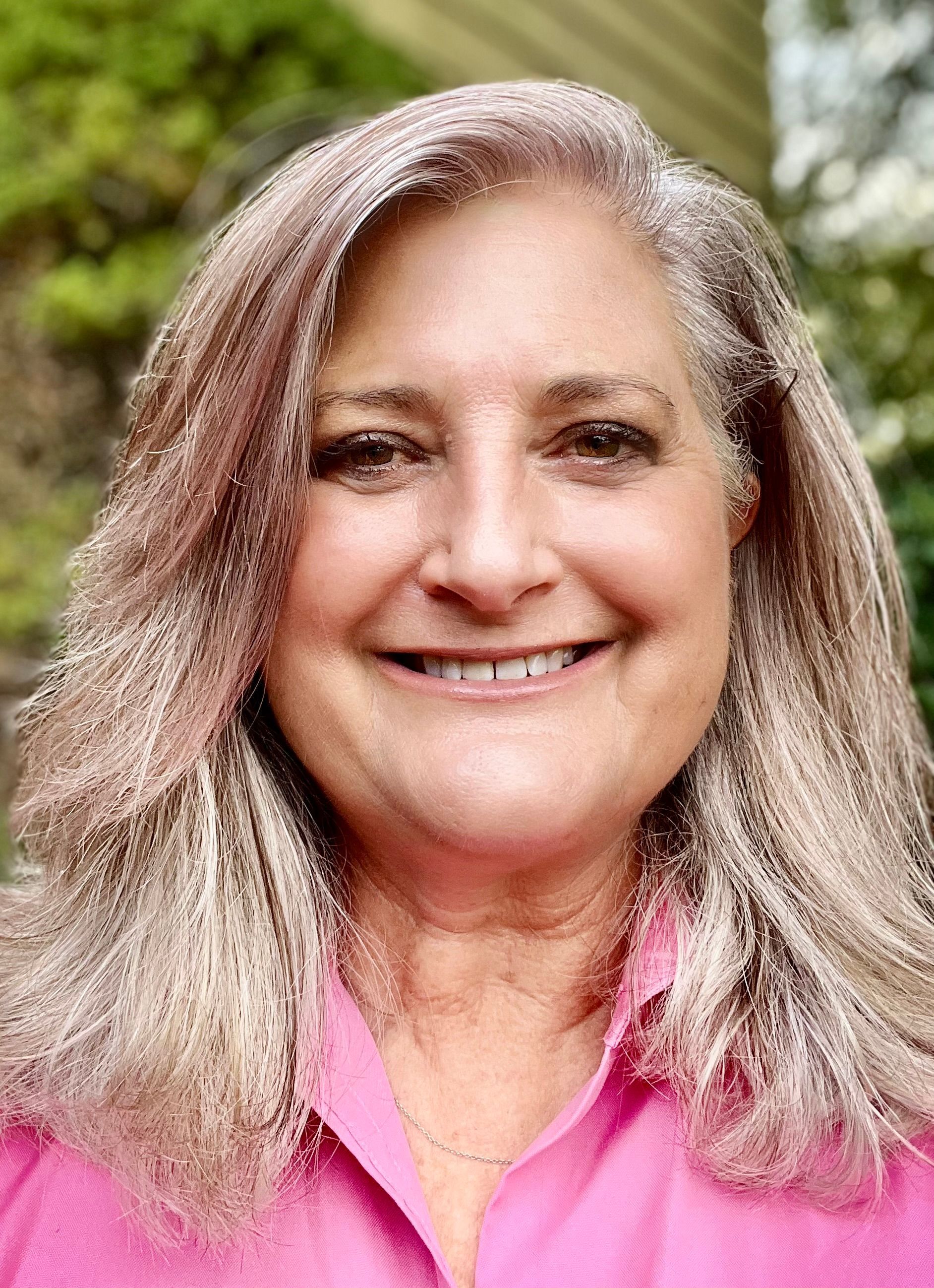 Woman with long, graying hair smiles, wearing a pink shirt, outdoors.