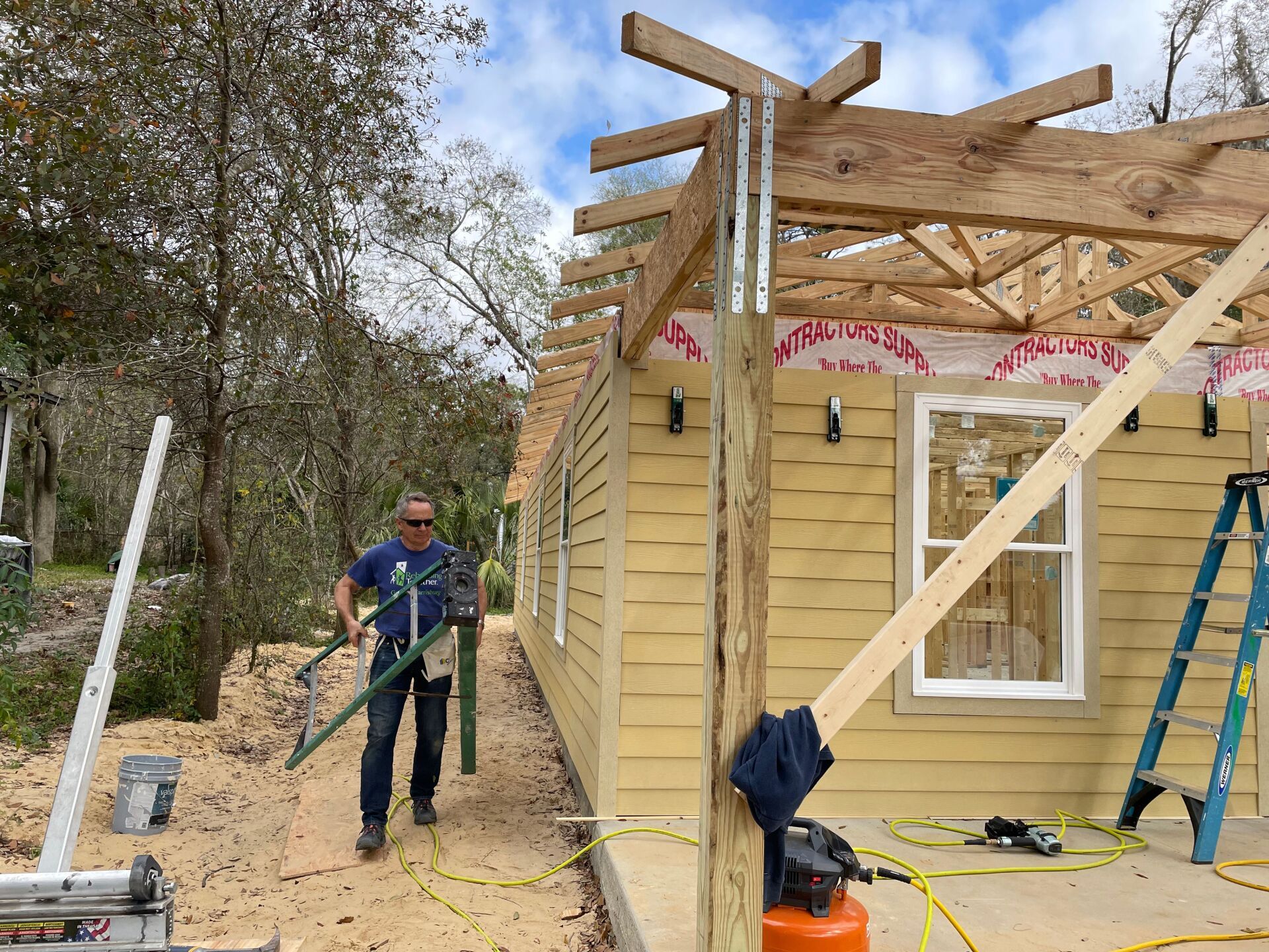 Man carrying metal poles at a building construction site. Yellow siding, wooden roof framing, blue sky.