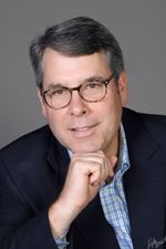 Man in glasses, navy jacket, hand on chin, smiles at the camera.