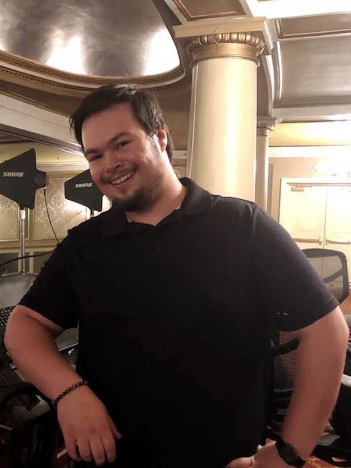Man in black polo shirt smiling, leaning on a desk indoors, near a white column.