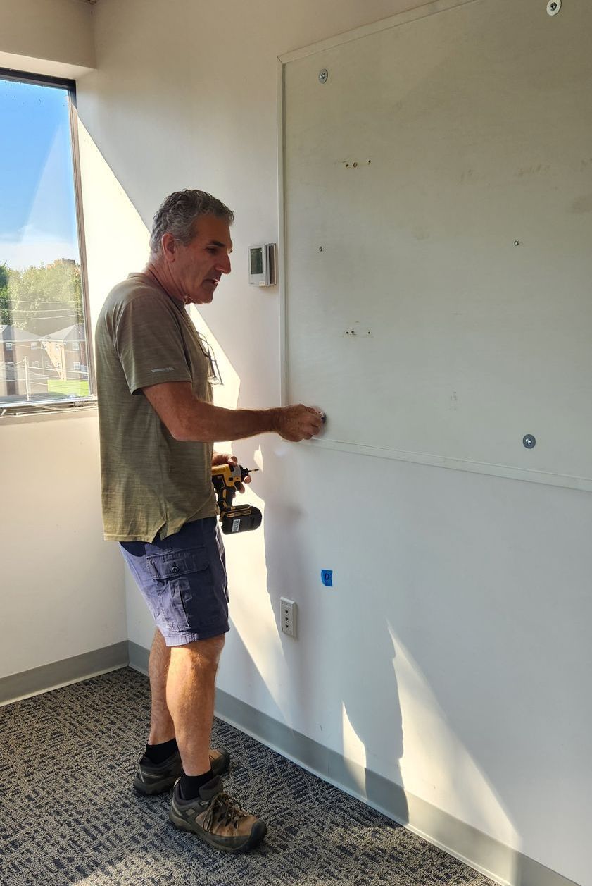 Man in shorts using a drill on a white wall in a room with a window.