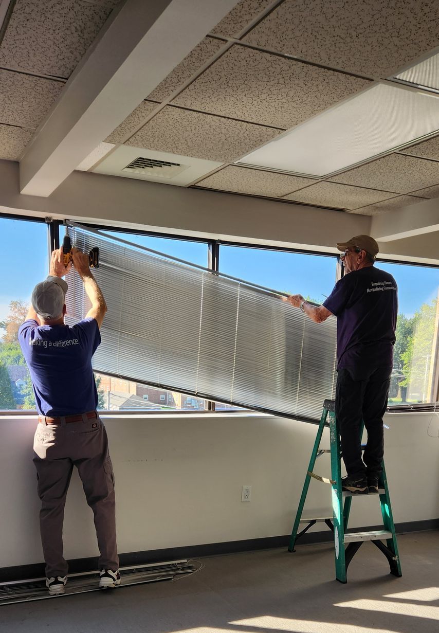 Two men installing a large, dark screen in front of a window inside a building.